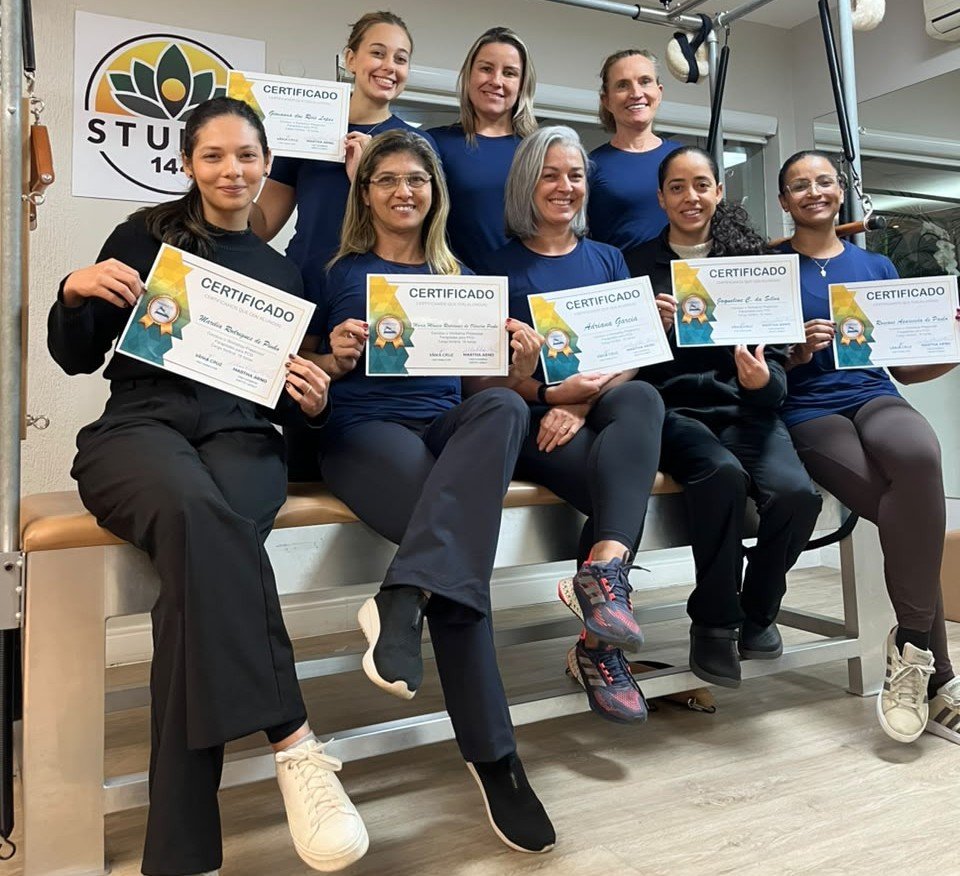 Participantes do workshop de fevereiro de 2024 sorrindo e segurando seus certificados, sentados em um aparelho de Pilates.
